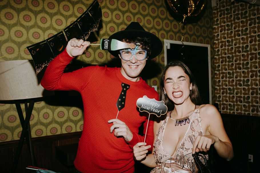 Two people posing at a party with photo booth props, wearing festive attire in front of patterned wallpaper.