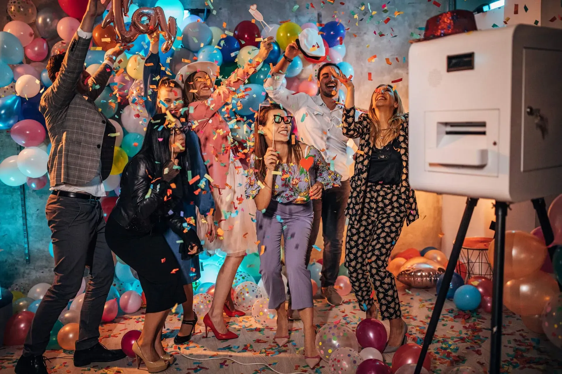 A group of friends celebrates in a room filled with colorful balloons and confetti near a photo booth.
