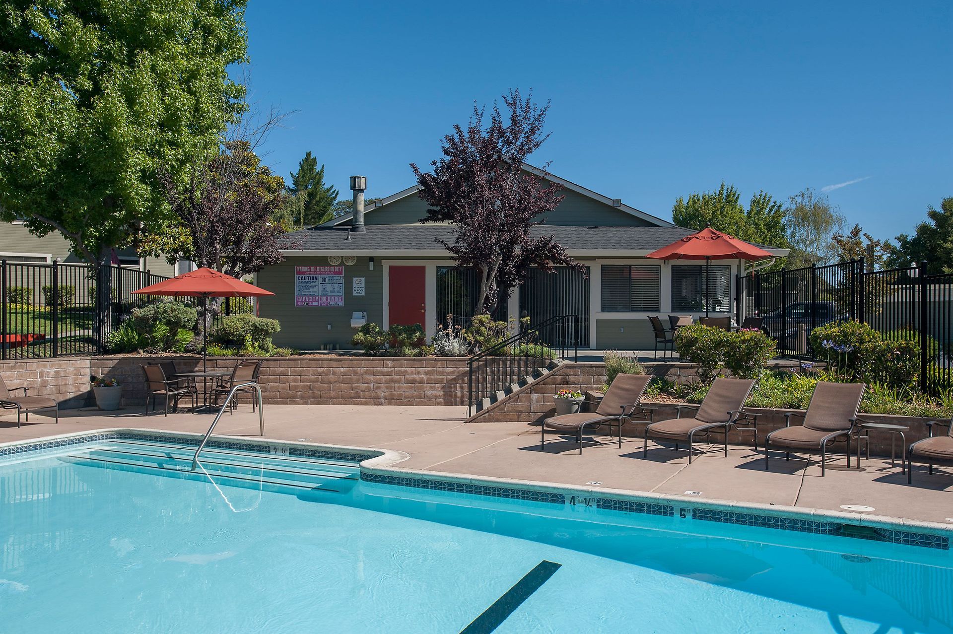 Outdoor pool area with lounge chairs, umbrellas, and a clubhouse in the background.