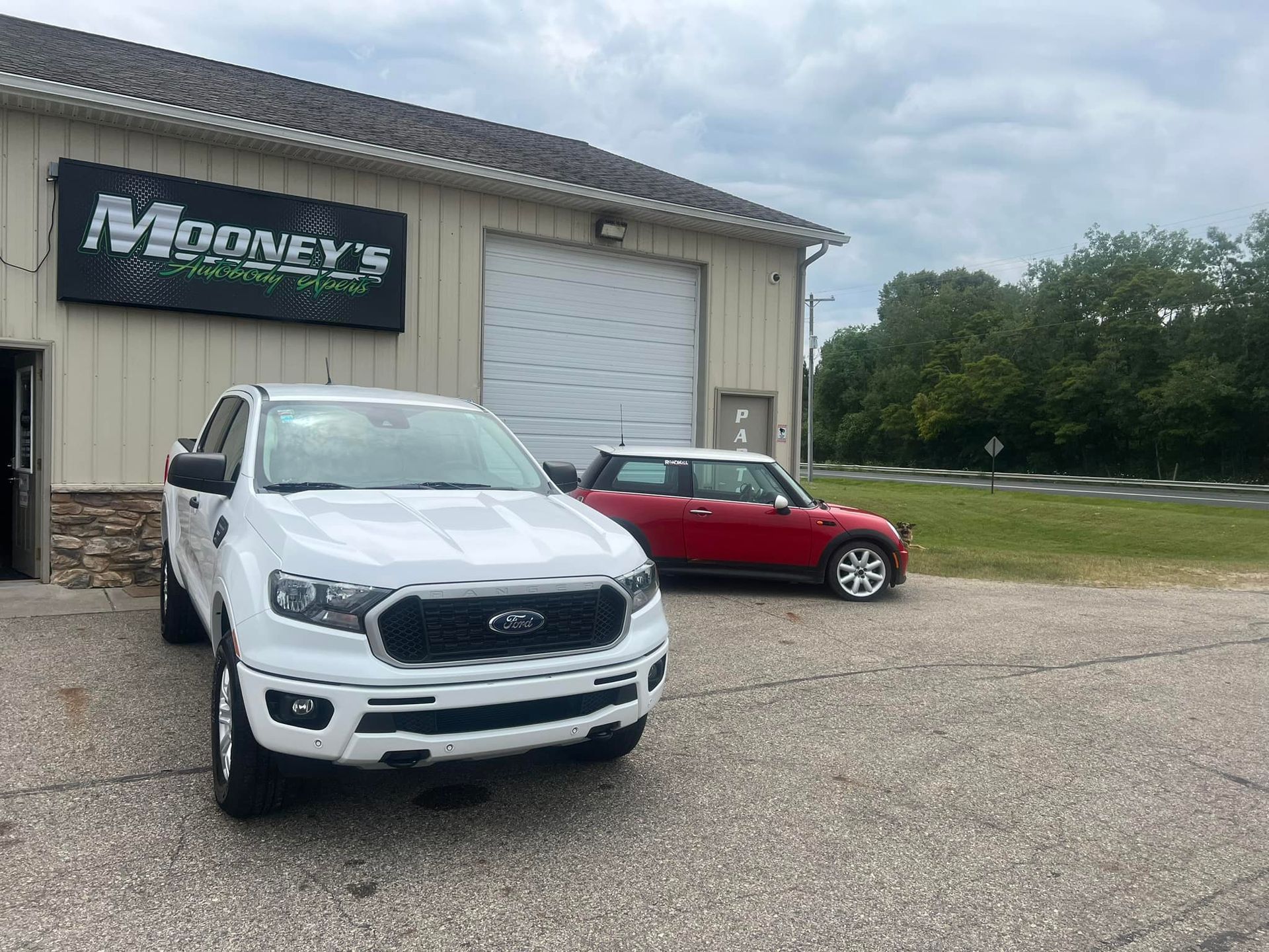 A white Ford Ranger pickup truck parked in front of an auto shop, with a small red car parked behind it.