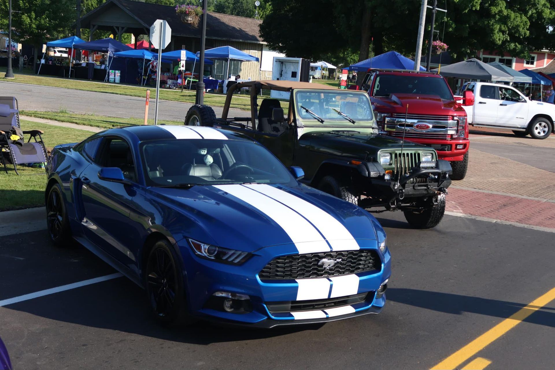 A blue Ford Mustang with white racing stripes parked next to an olive green Jeep at an outdoor car show.
