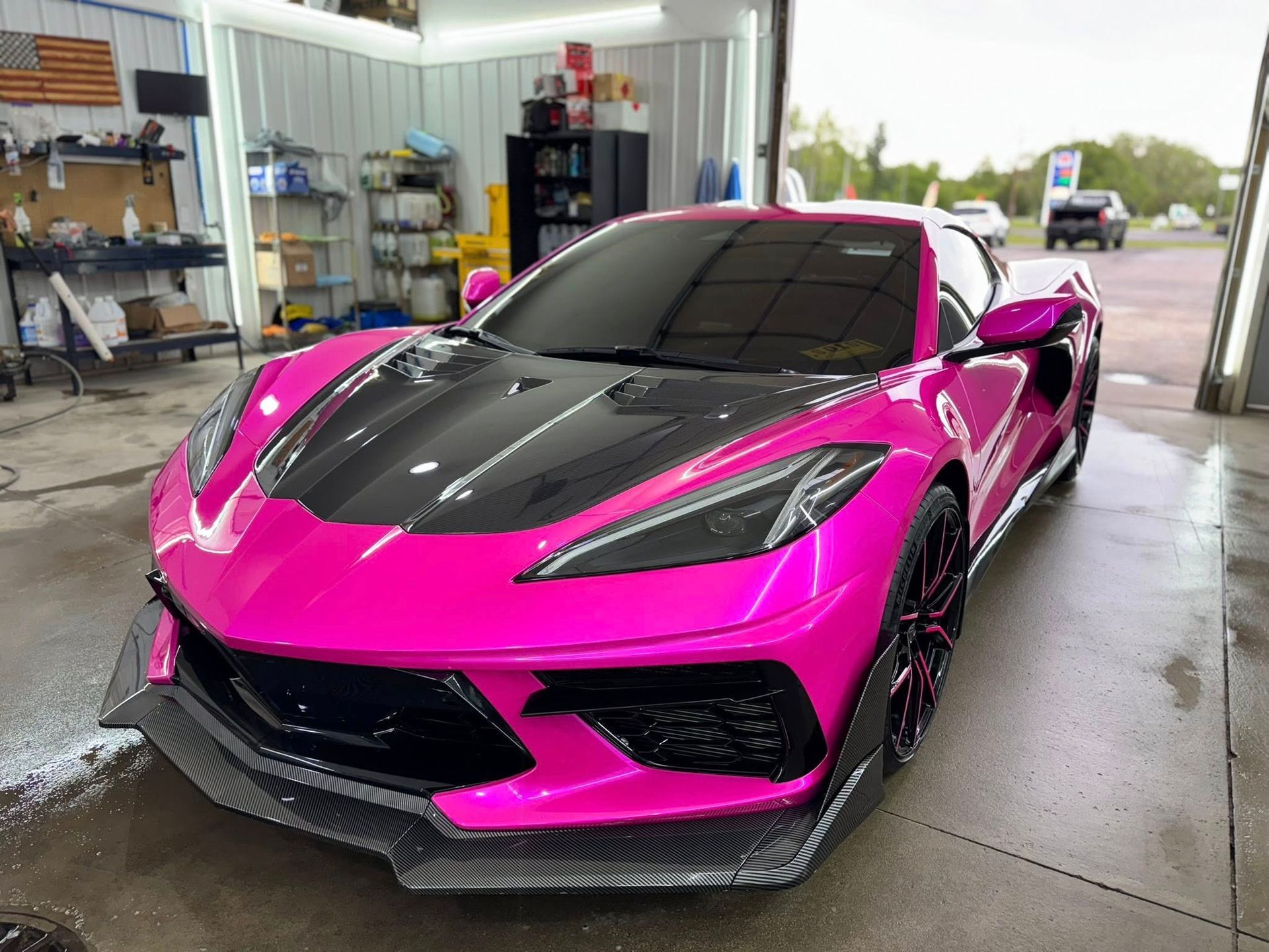 A bright pink Chevrolet Corvette with a carbon fiber hood and front splitter parked inside a workshop.