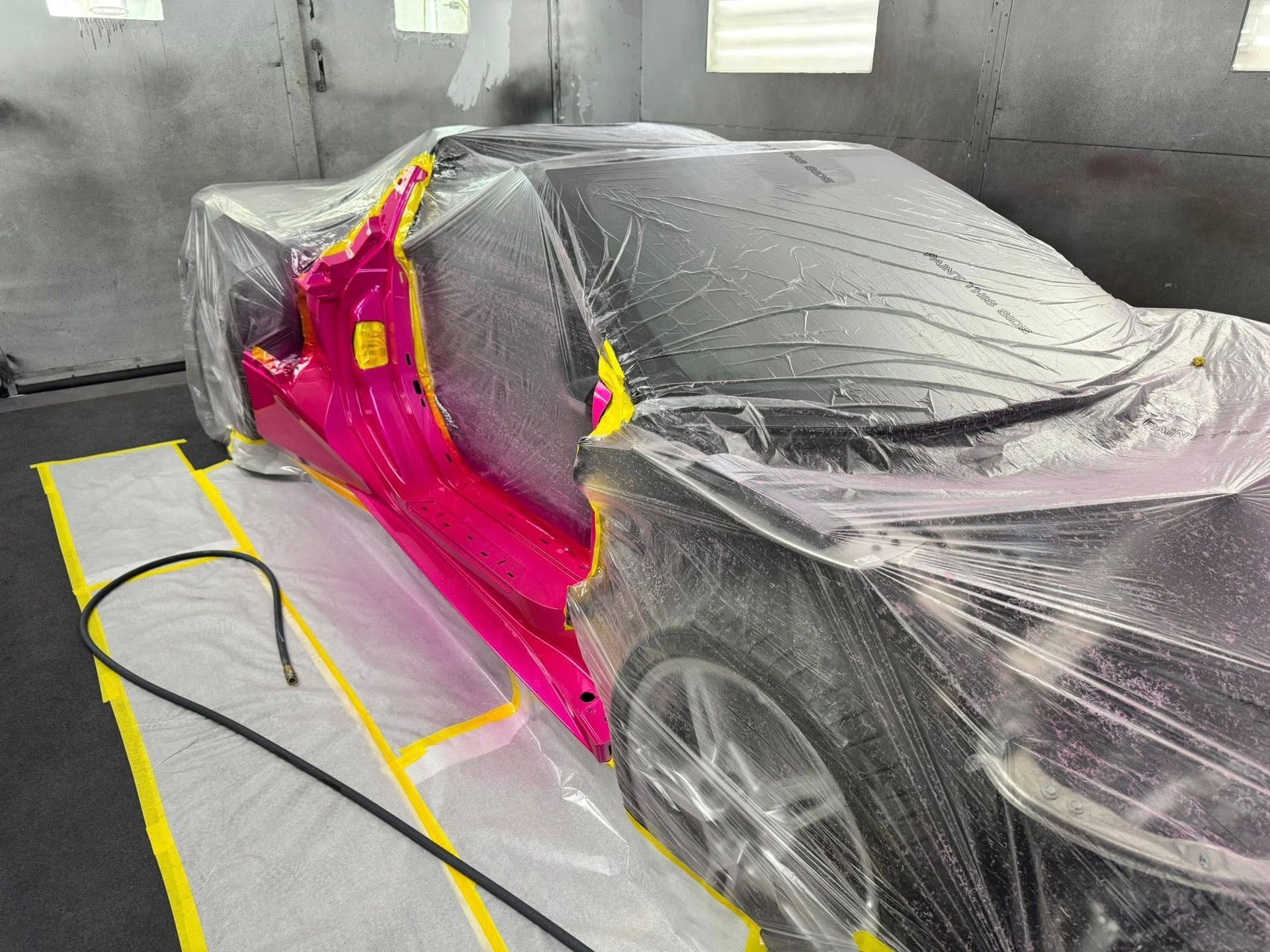 A sports car partially covered in plastic sheeting inside a spray booth, with a vibrant pink side panel exposed.