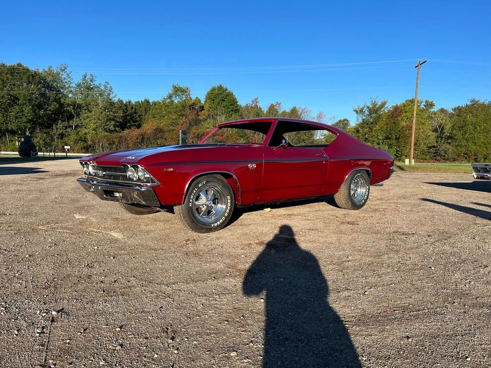 A shiny, deep red classic muscle car parked on a gravel lot under a clear blue sky.