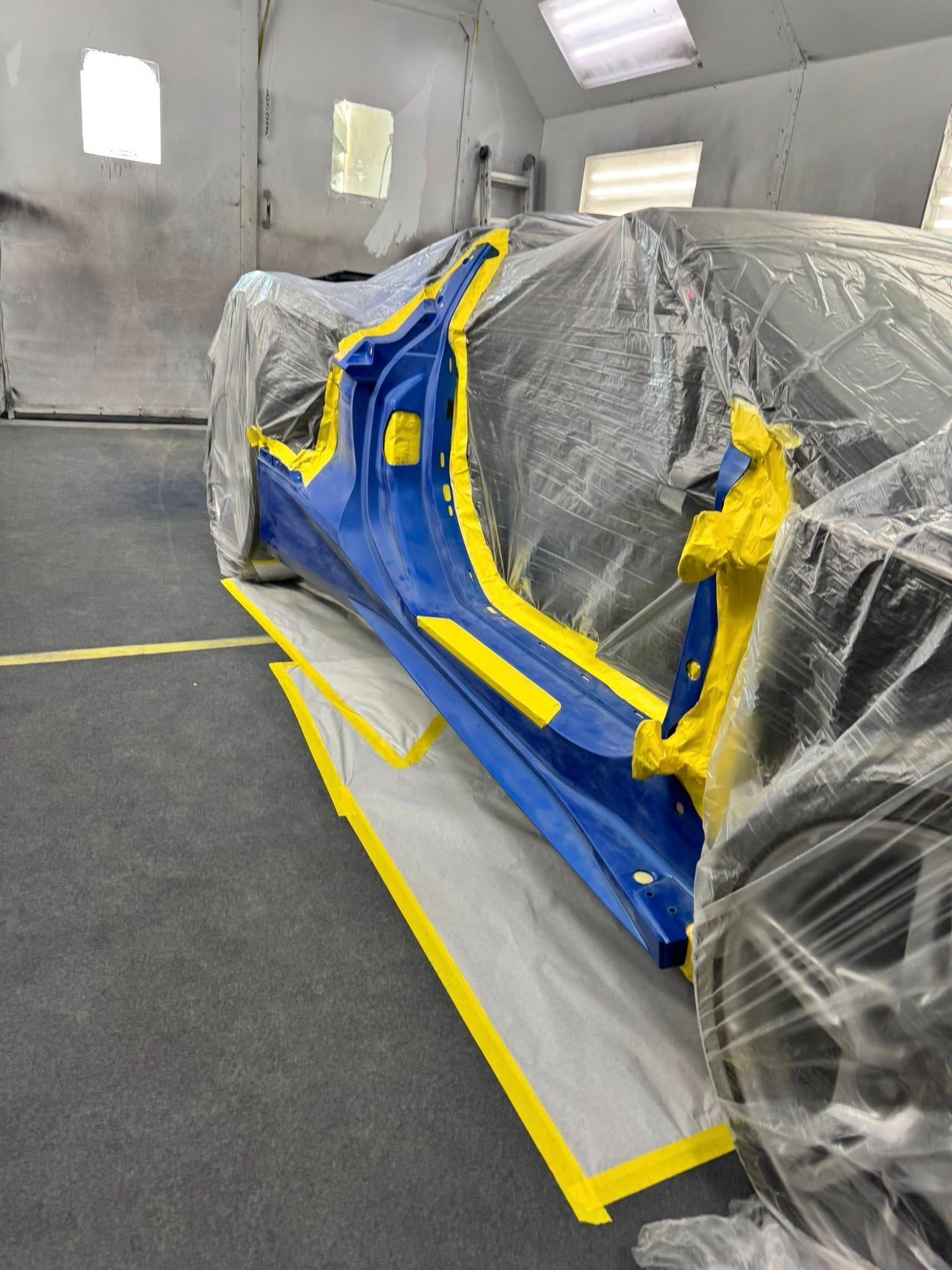 A side panel of a blue car, masked with yellow tape and plastic wrap inside an auto body paint booth.