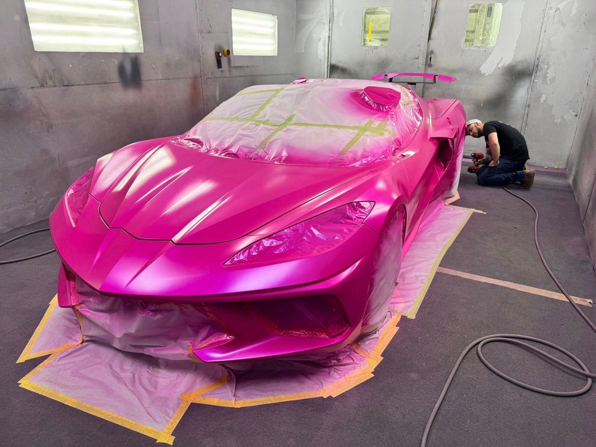 A technician spray-paints a bright pink sports car inside a paint booth.