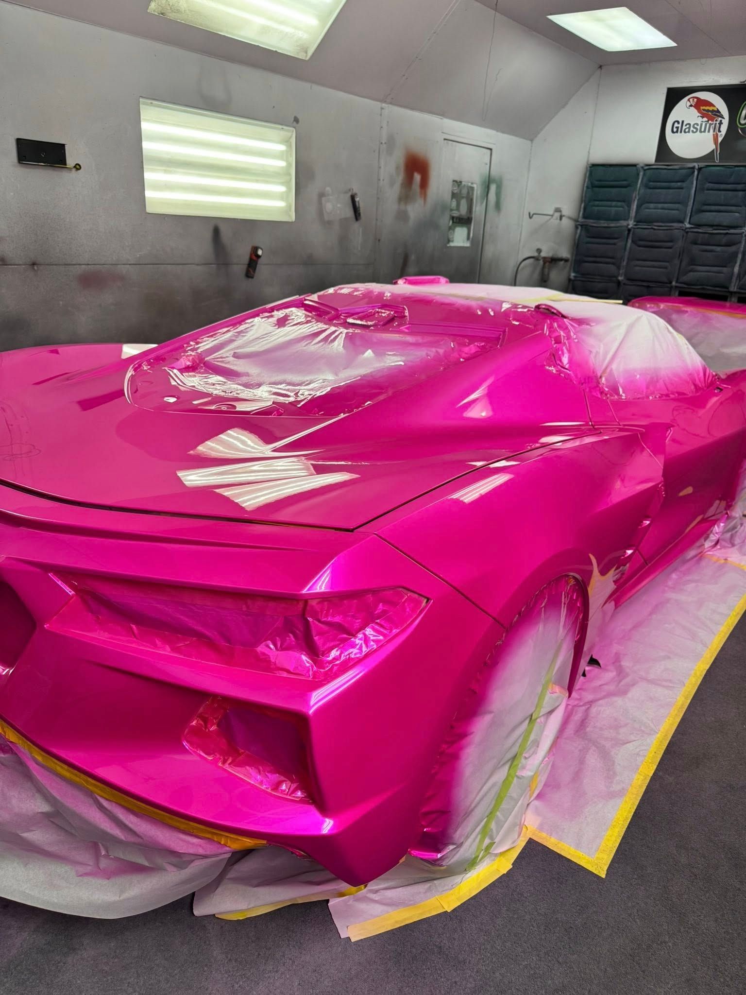 A bright pink, glossy sports car parked in an auto body shop, partially covered with protective masking tape and plastic.