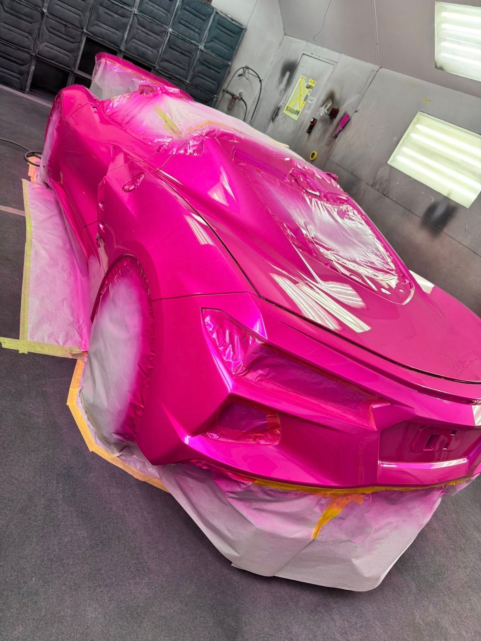 A bright pink sports car sits in a paint booth, partially covered in protective plastic sheeting.