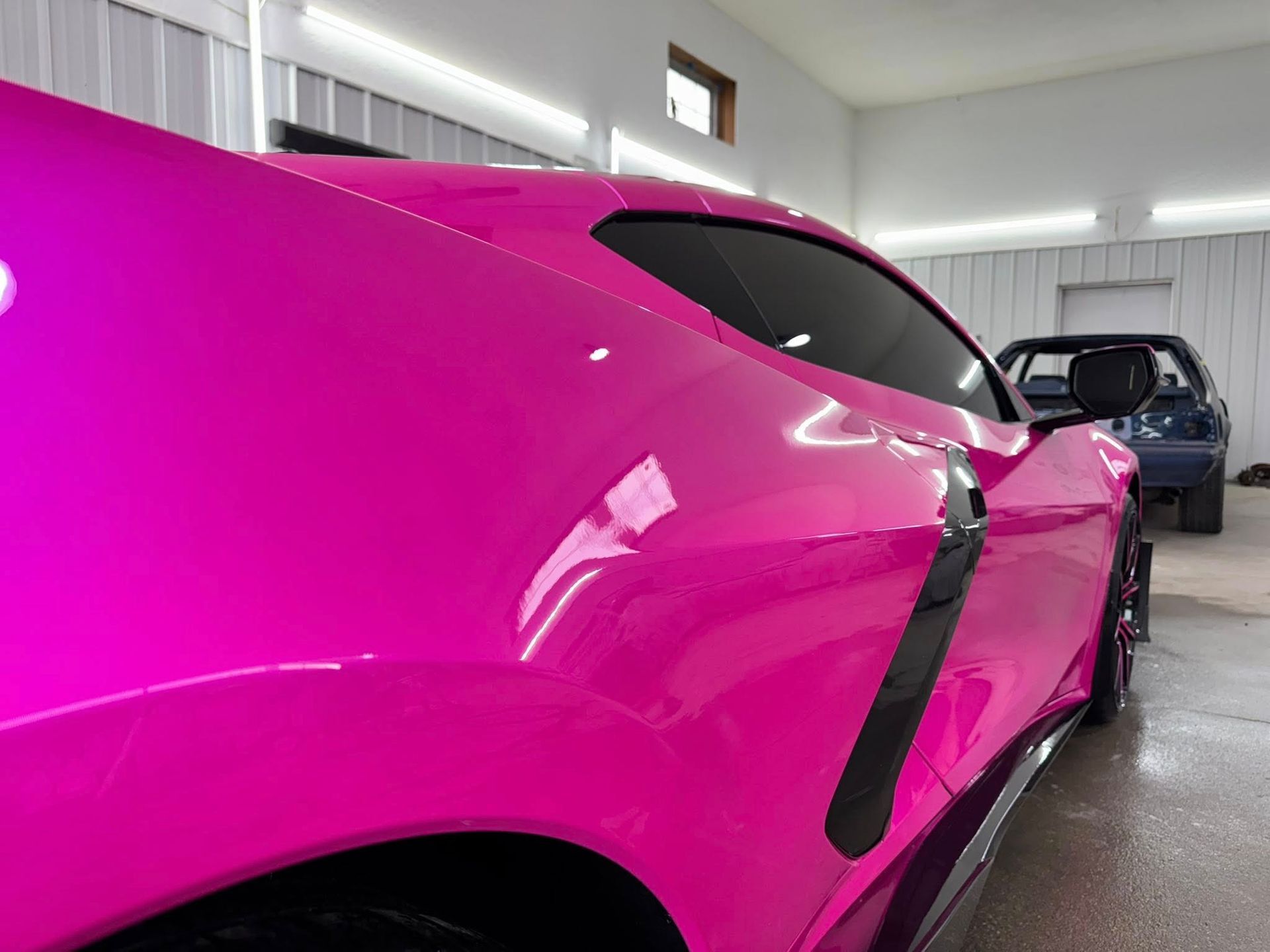 A glossy magenta Chevrolet Corvette parked in a bright, modern garage with a dark car in the background.