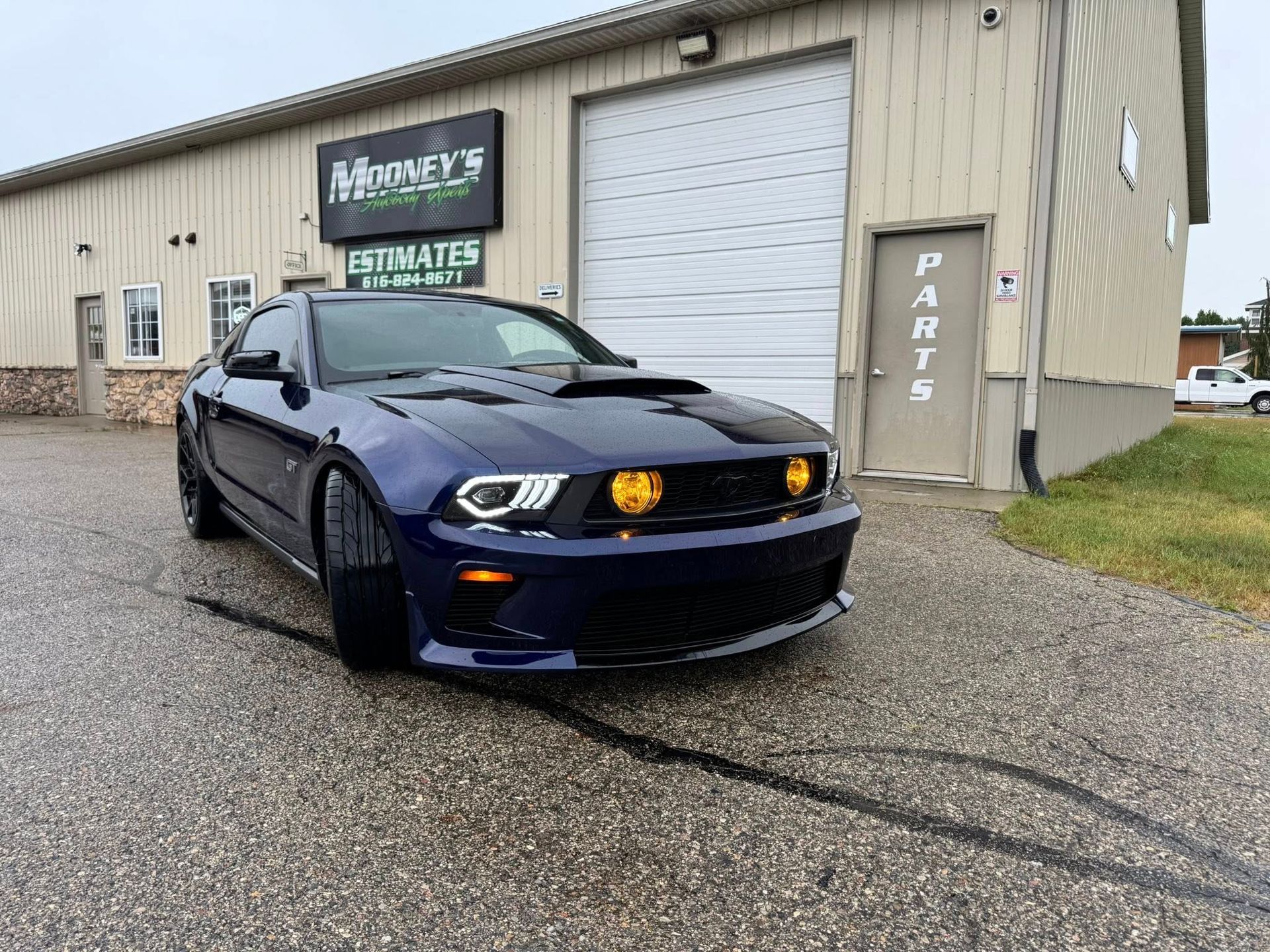 A dark blue Ford Mustang parked on a gravel lot in front of an auto repair shop with a 
