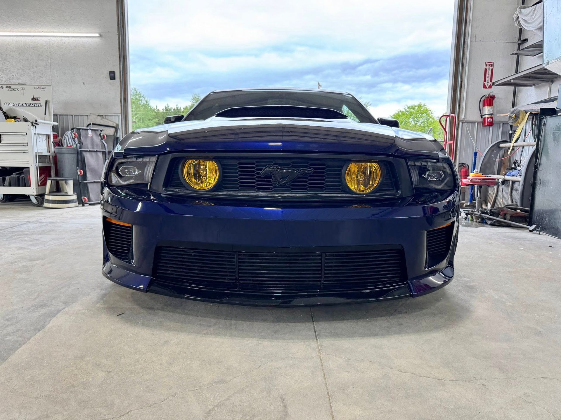 A blue Ford Mustang with yellow fog lights parked inside a garage.