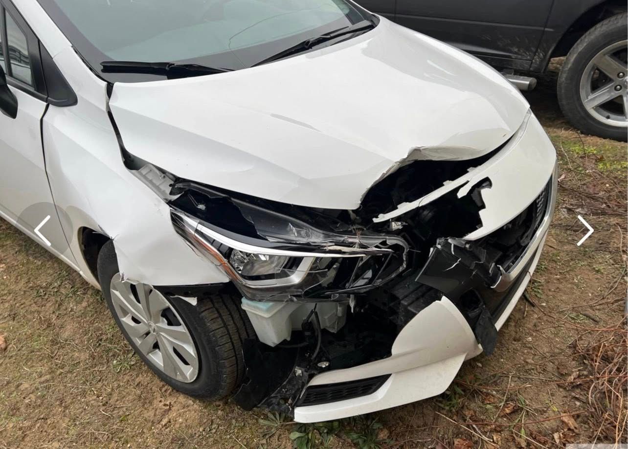 A white car with significant front-end damage, including a crushed hood, bumper, and headlight, parked on dirt.