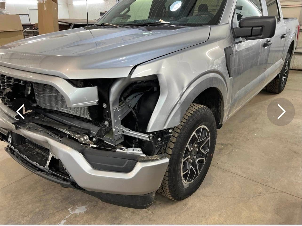 A silver pickup truck parked indoors with significant front-end collision damage, including a missing headlight.