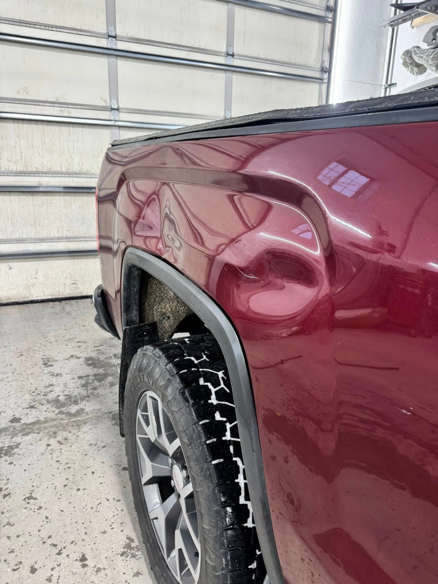 A side view of a maroon pickup truck bed showing a noticeable dent above the rear wheel arch inside a garage.