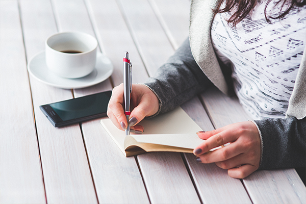 a woman is writing in a notebook next to a cup of coffee and a cell phone .