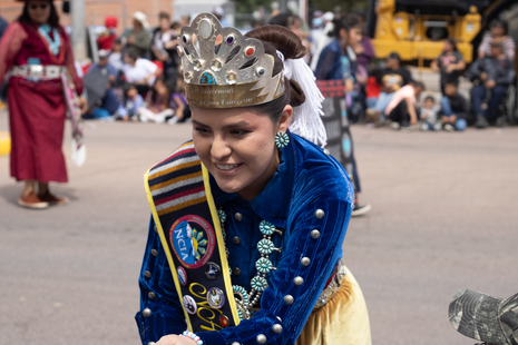 A woman wearing a crown and sash is smiling in a parade.