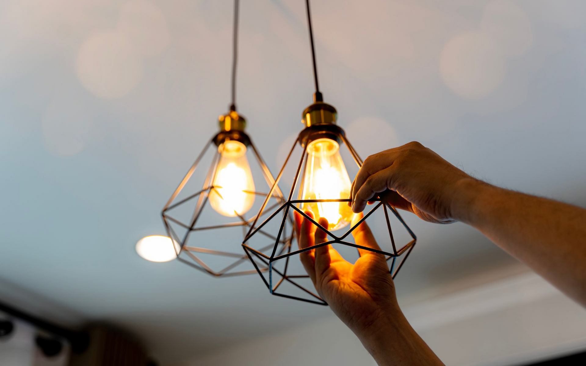 Person's hands adjusting a lit light bulb in a geometric pendant lamp hanging from a white ceiling.