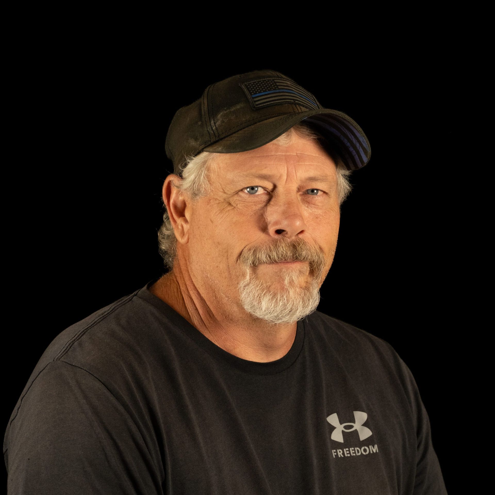 Man with graying beard and baseball cap, wearing a black shirt, looking at the camera against a black background.