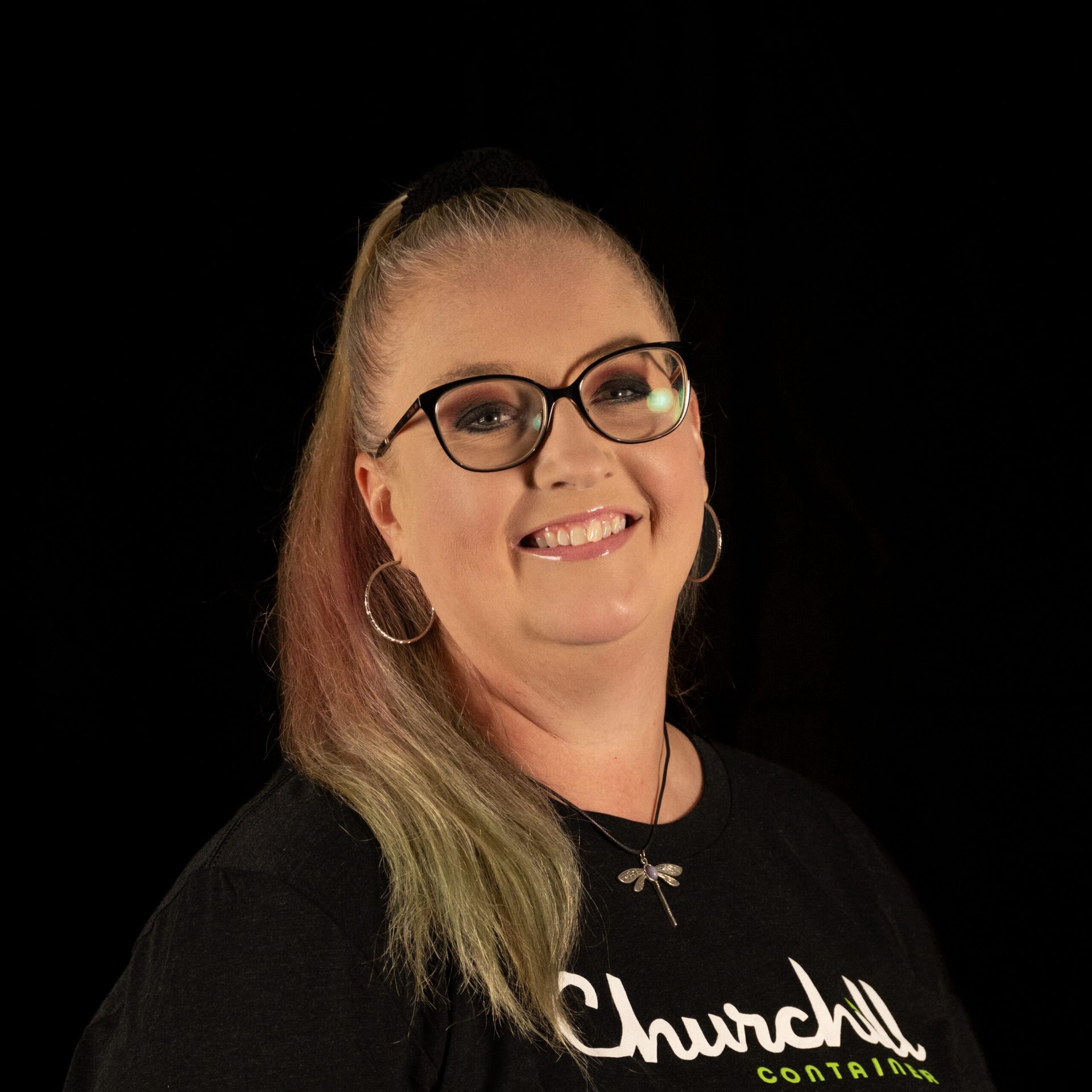 Woman wearing glasses, smiling, with a high ponytail and a black shirt that says 