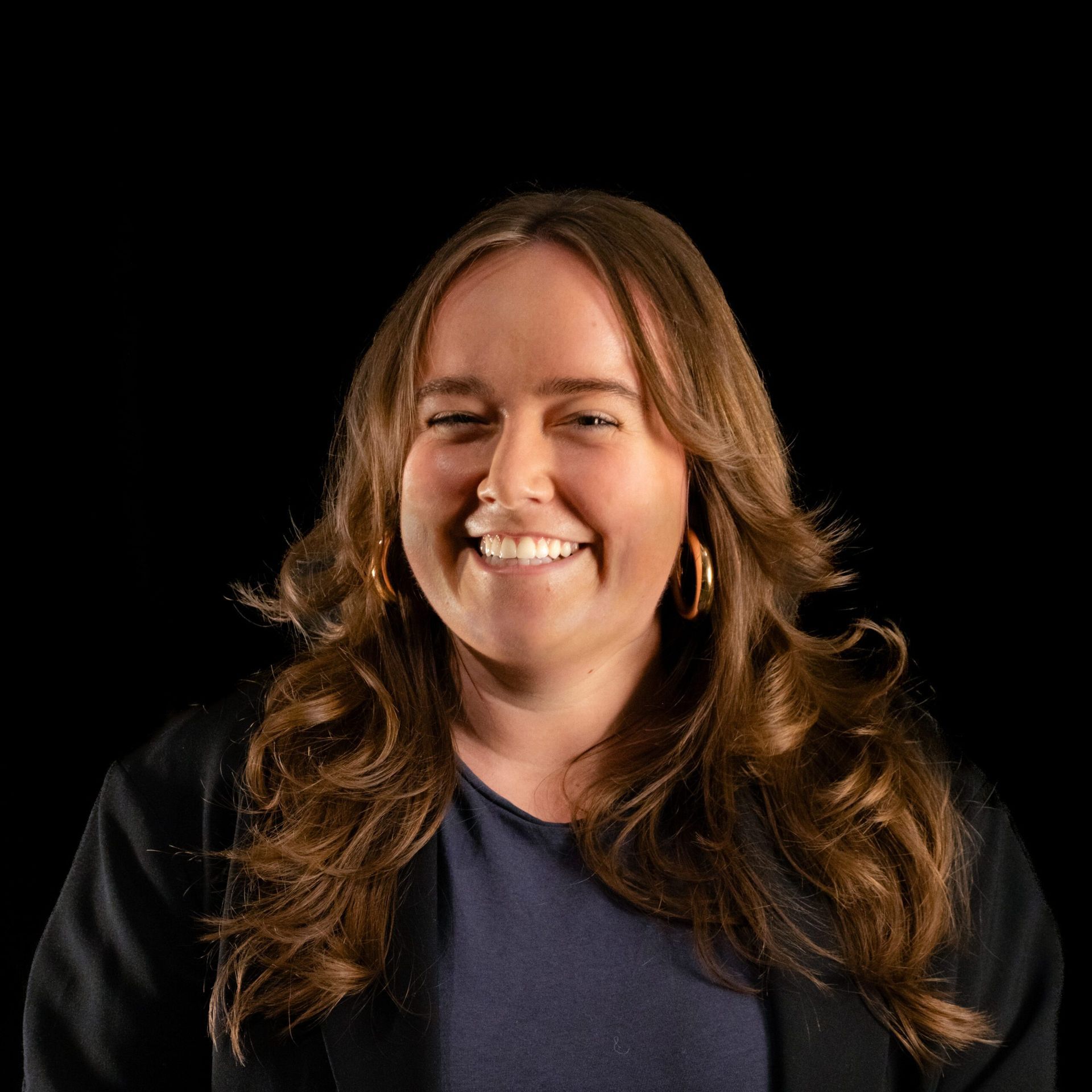 Woman with long brown hair smiles broadly against a black backdrop, wearing a dark blazer and hoop earrings.