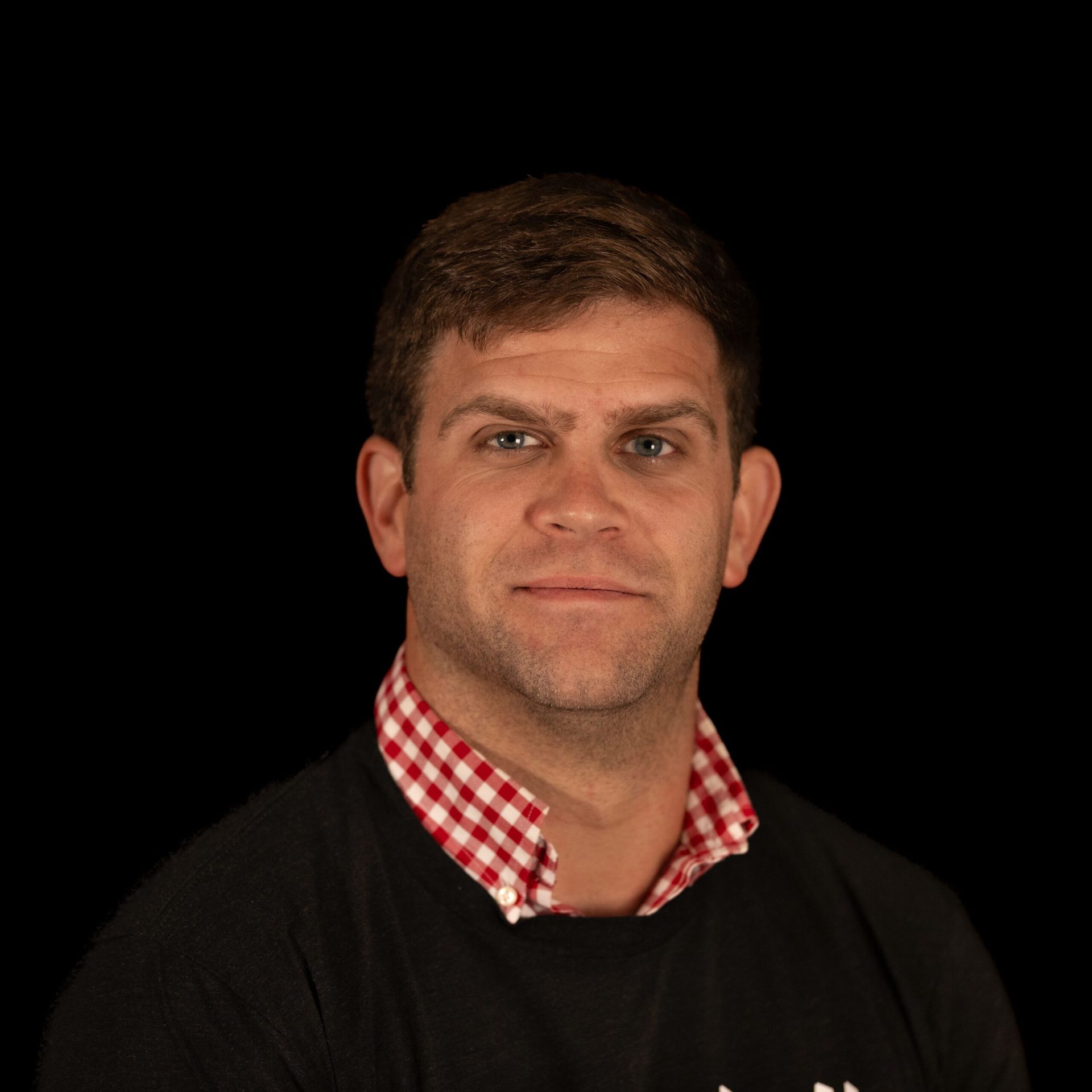 Man with short brown hair wearing a black shirt over a red and white checkered collared shirt, against a black background.