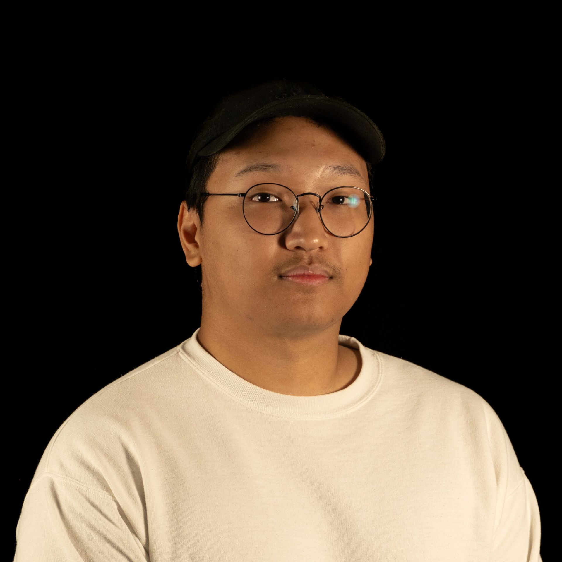 Man wearing glasses and a black cap, looking at the camera. He is wearing a white crew neck shirt against a black background.