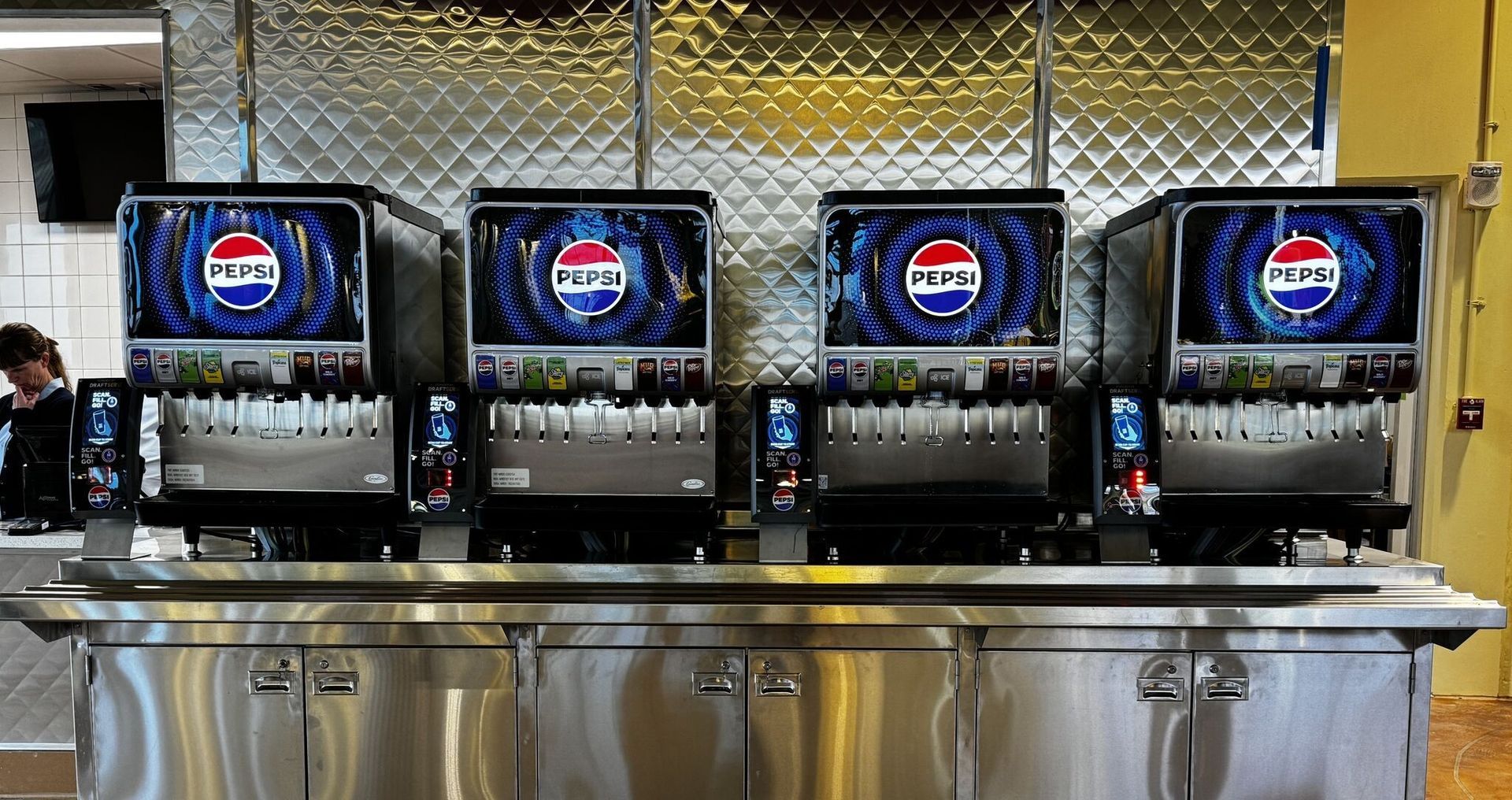 Four Pepsi soda fountain machines with digital screens, stainless steel counter, and a person in the background.