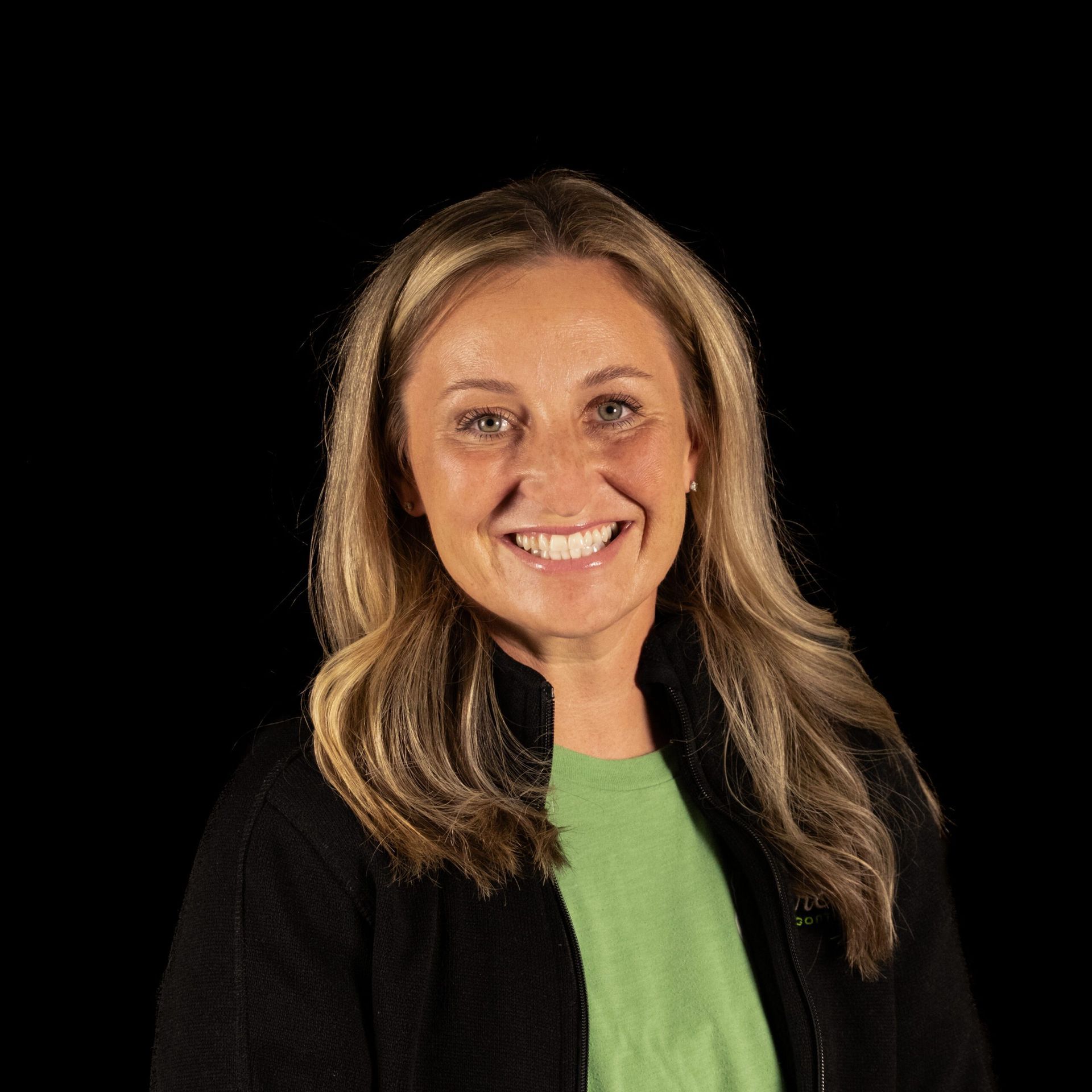 Smiling woman in a black jacket and green shirt against a black background.