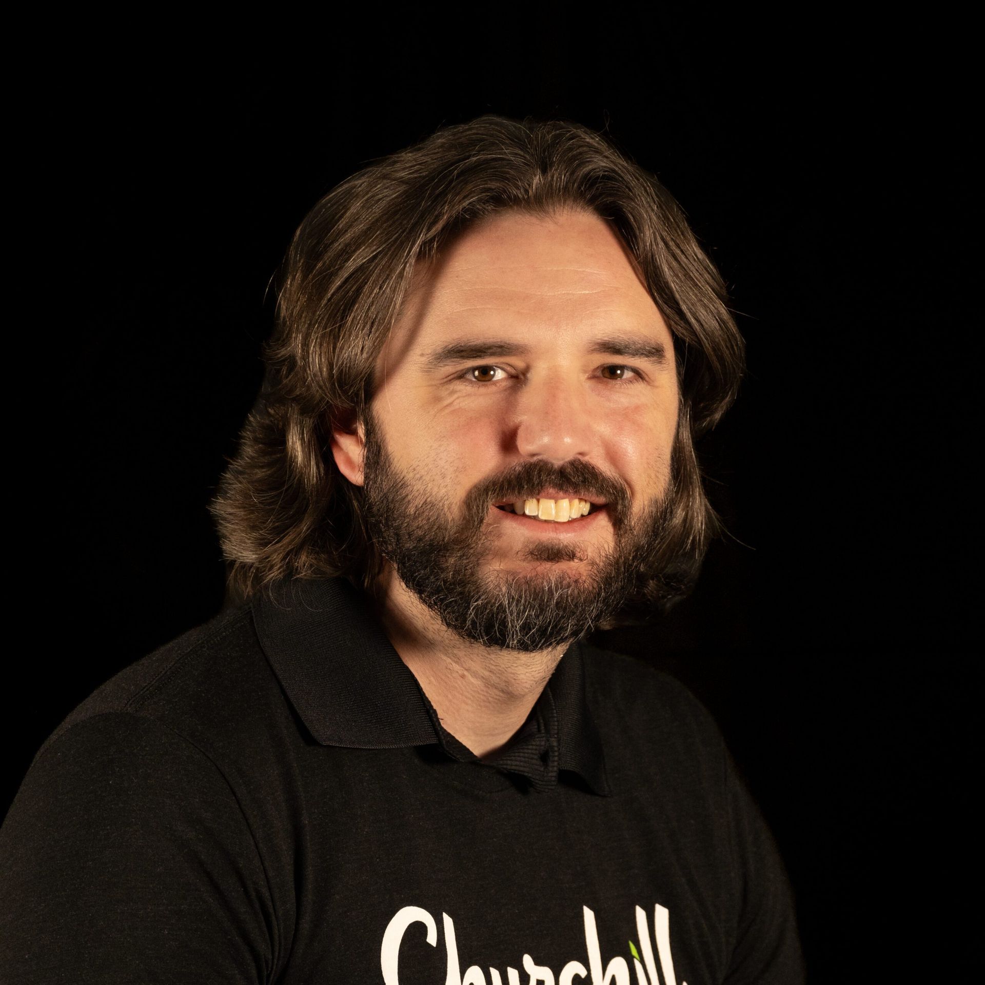 Man with brown hair and beard, wearing a black shirt with white text, smiles against a black background.