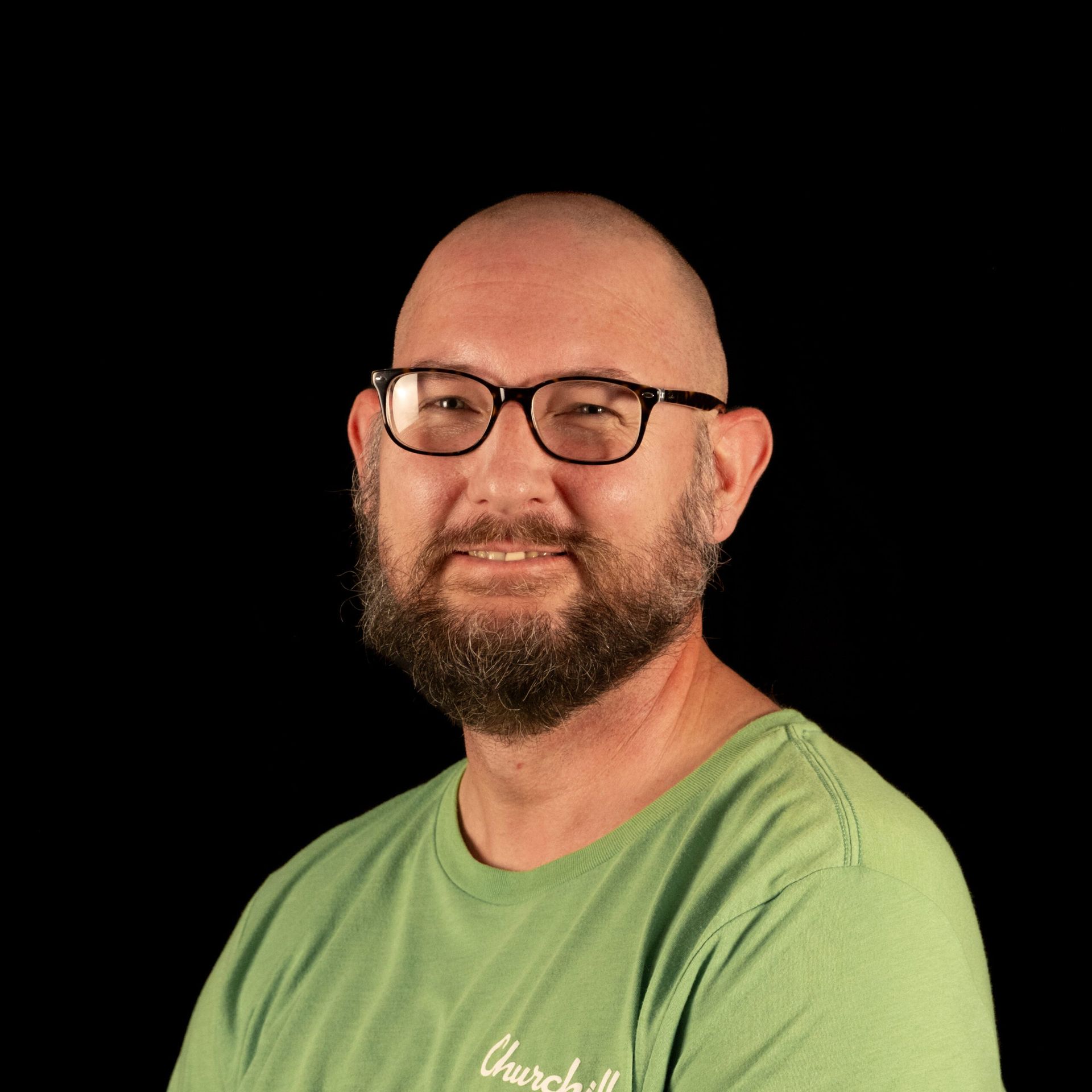 Man with a beard and glasses smiles, wearing a green shirt, against a black background.