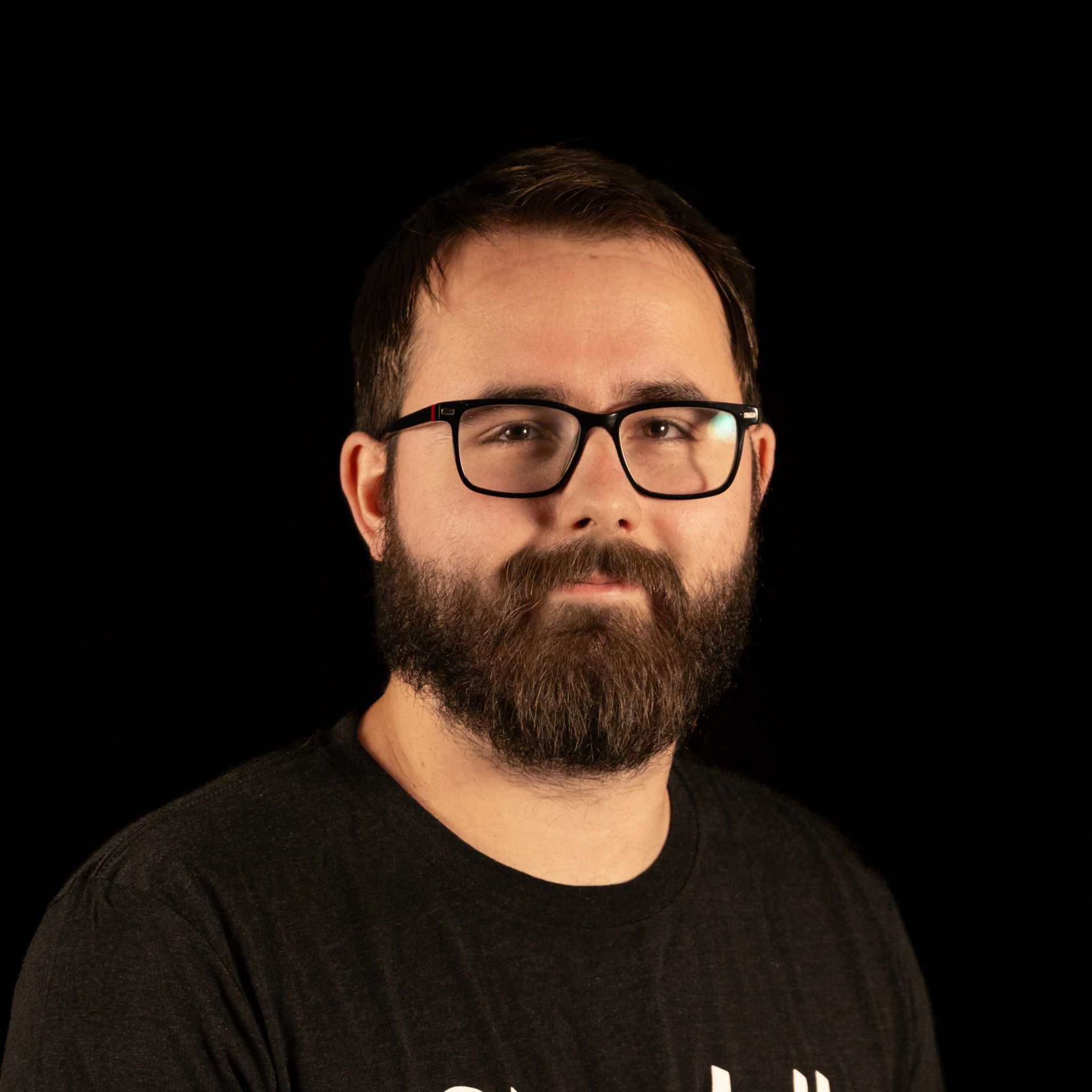 Man with glasses and beard against a black background. He is wearing a dark t-shirt and smiling slightly.