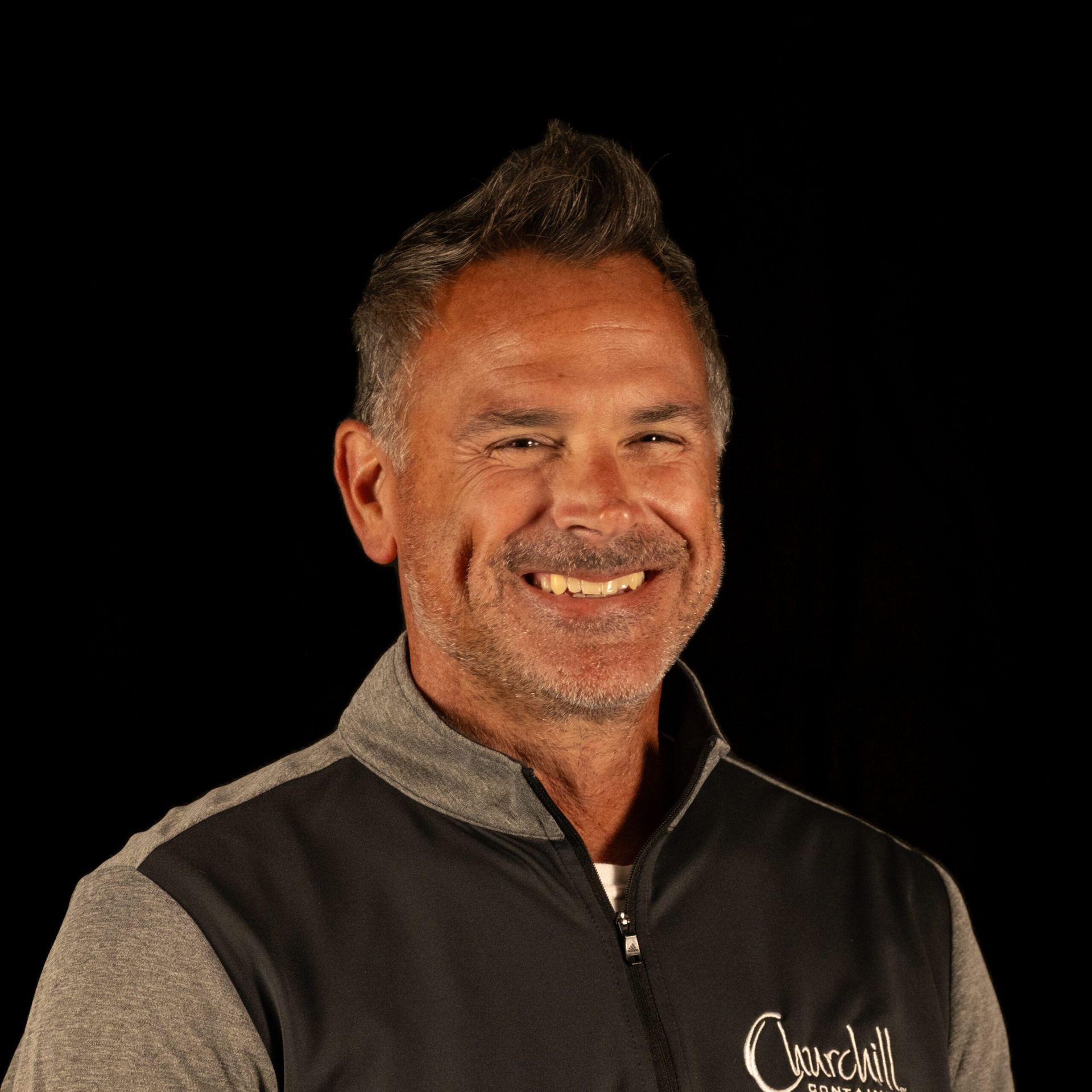 Man smiling, wearing a gray and black vest, against a black background.