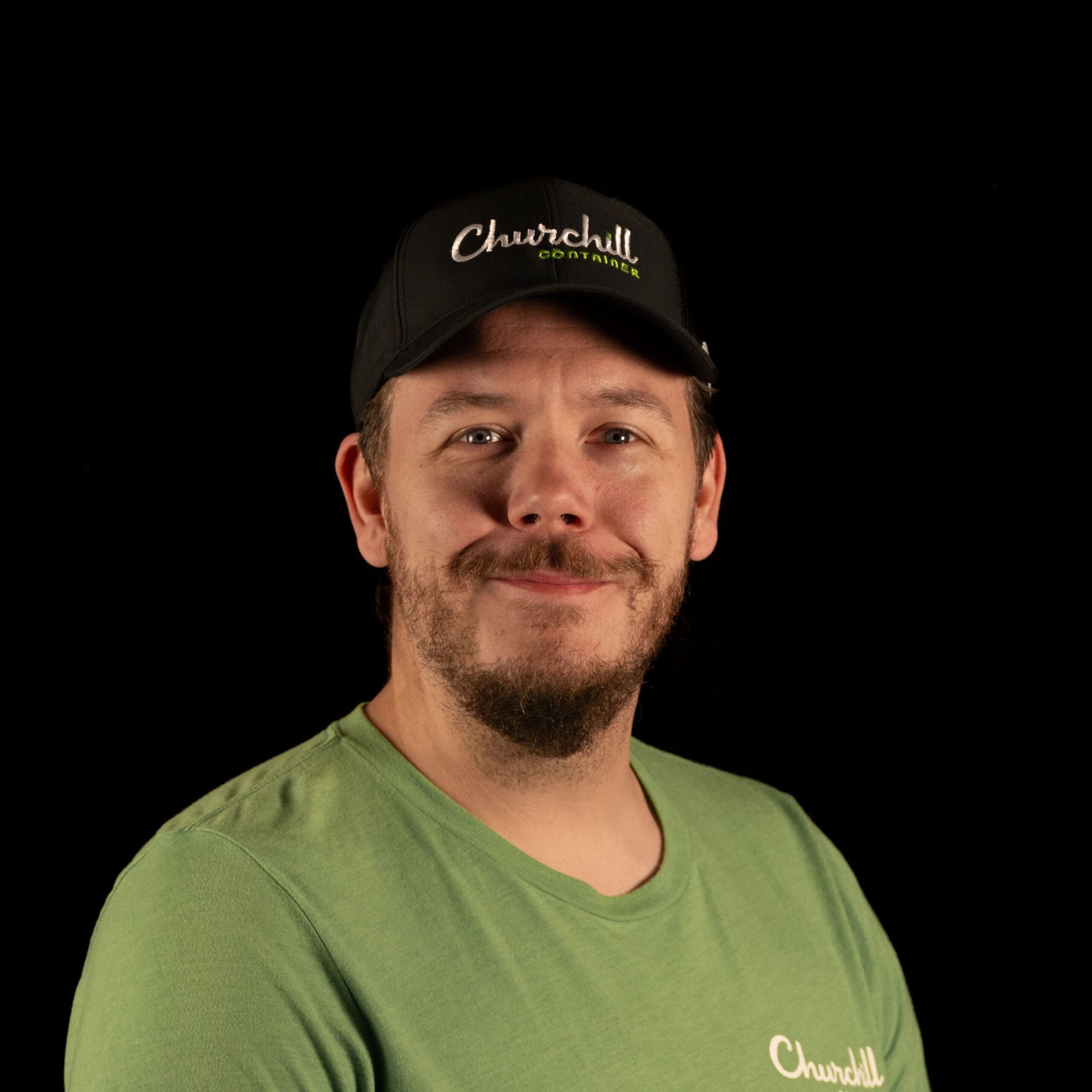 Man wearing a Churchill Coffee cap and green shirt smiles against a black background.