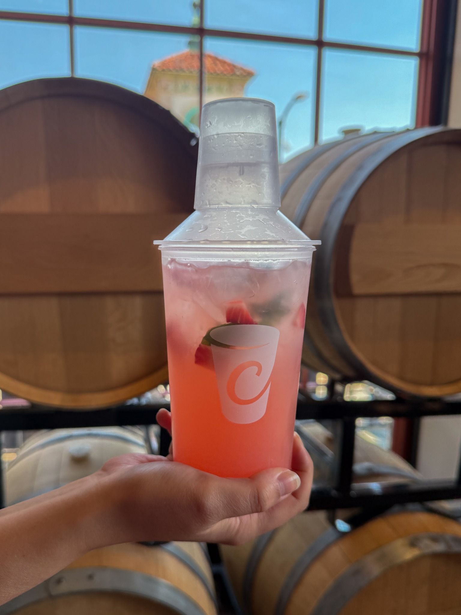 A person holding a pink drink in a clear cup with a lid on, near wooden barrels.