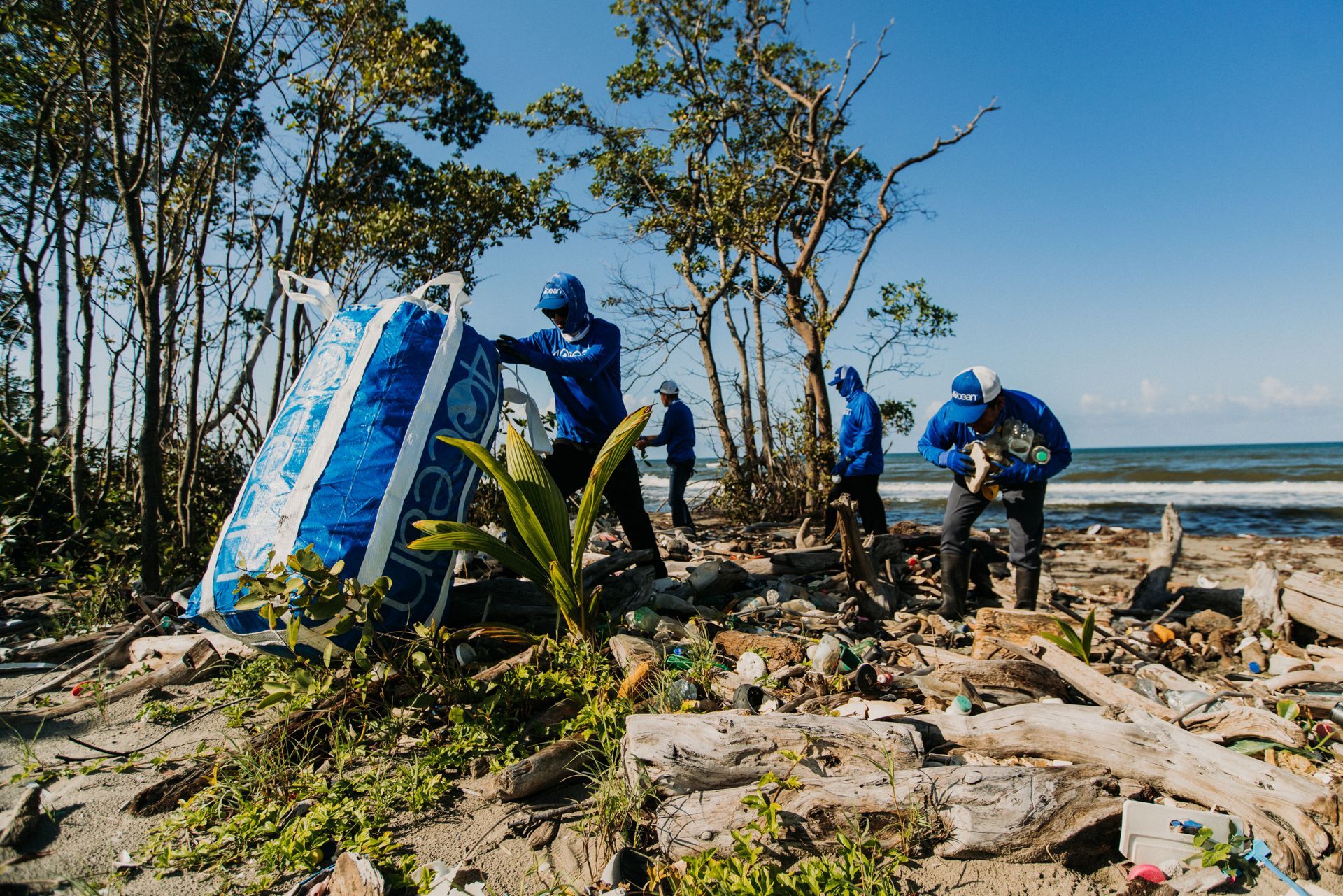 People in blue cleaning up trash on a beach, collecting a large blue and white object.