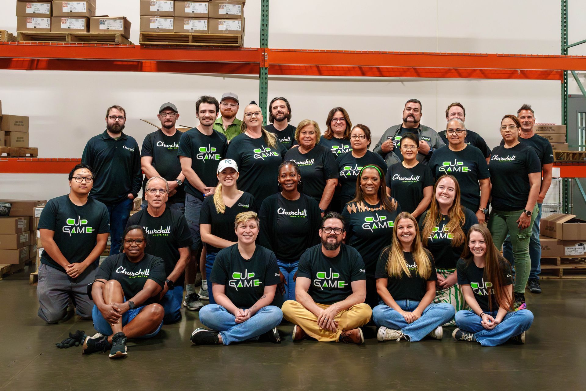 Group photo of people in matching shirts in a warehouse setting, smiling, many rows deep.