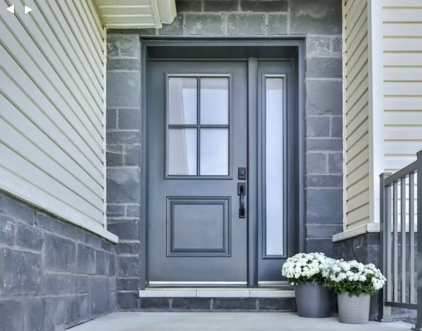 Porte d'entrée grise avec fenêtre latérale, entourée de briques grises et de bardage, fleurs blanches en pots.