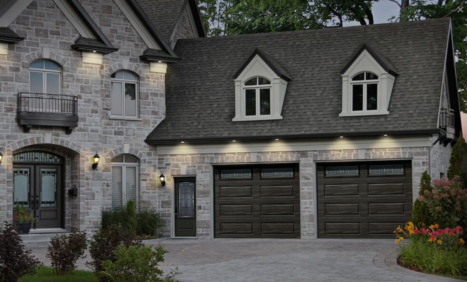 Maison en pierre avec garage pour deux voitures, portes de garage marron foncé et allée pavée.