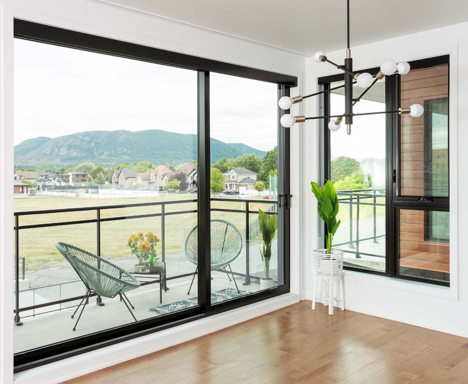 Bright room with large windows overlooking a balcony and landscape; modern chandelier, wood floors, and minimalist decor.