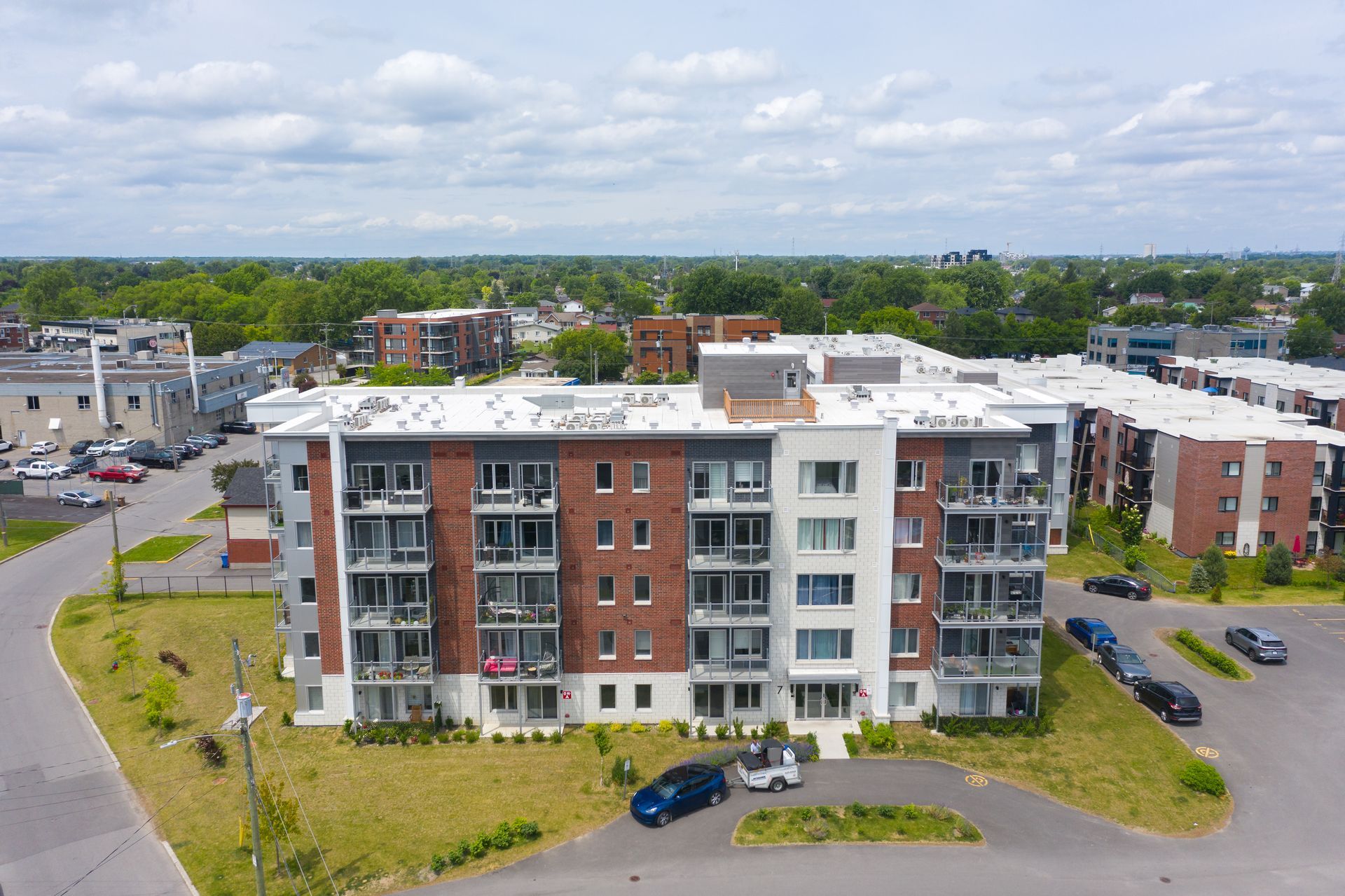 Immeuble d'appartements à plusieurs étages avec façade en briques rouges et blanche. Parking avec voitures au premier plan et arbres en arrière-plan.