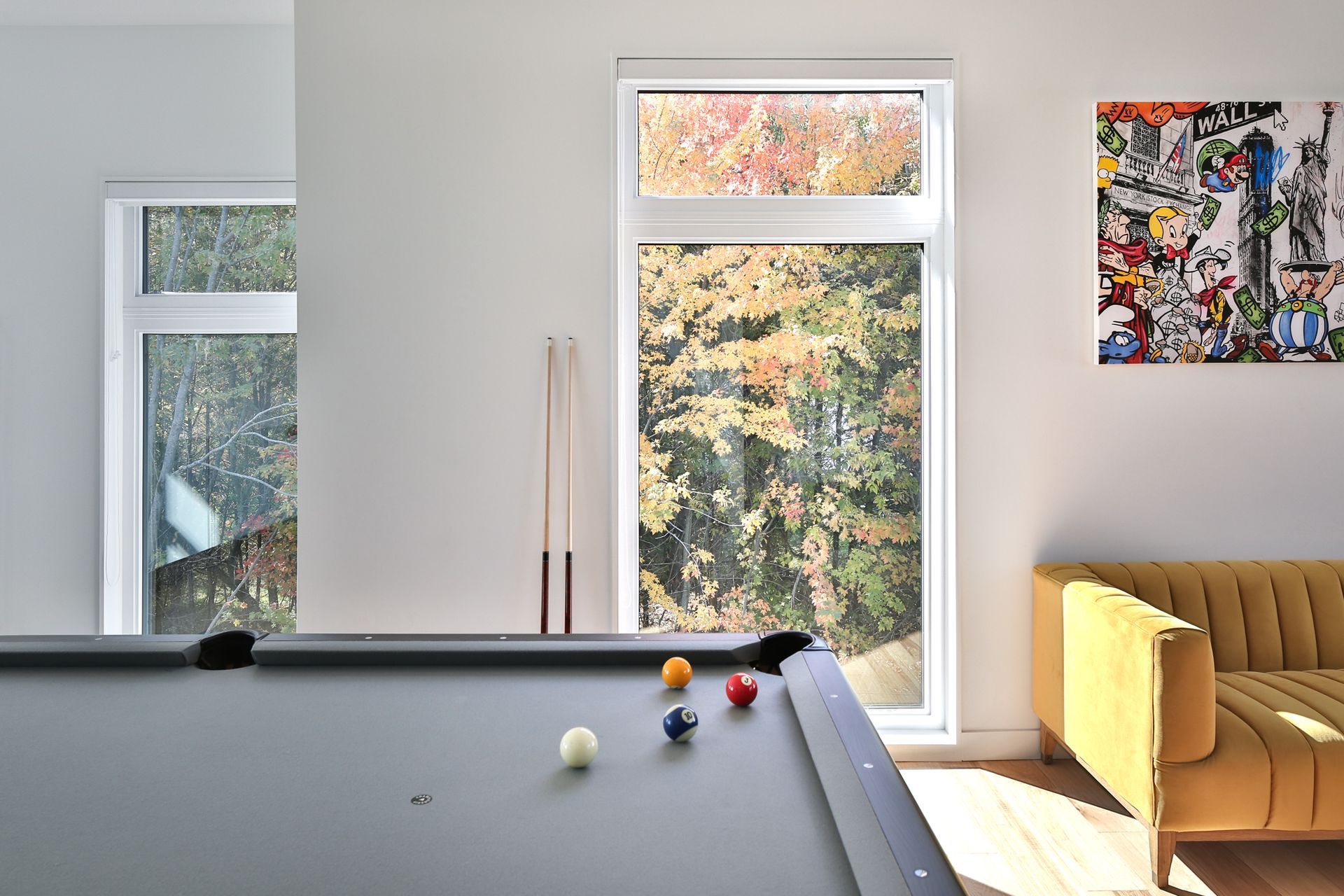 Table de billard dans une pièce avec fenêtres, boules colorées, canapé jaune, œuvre d'art sur le mur.