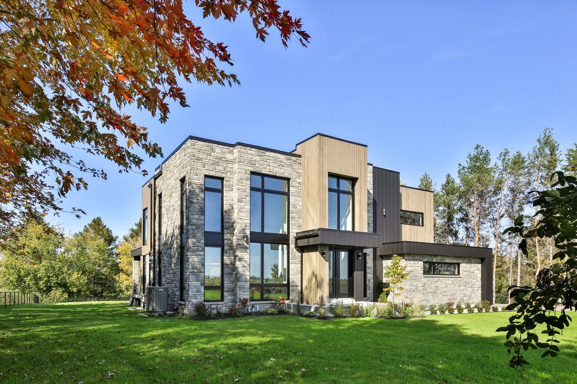 Maison moderne à deux étages avec extérieur en pierre et bois, grandes fenêtres, située sur une pelouse verte avec feuillage d'automne.