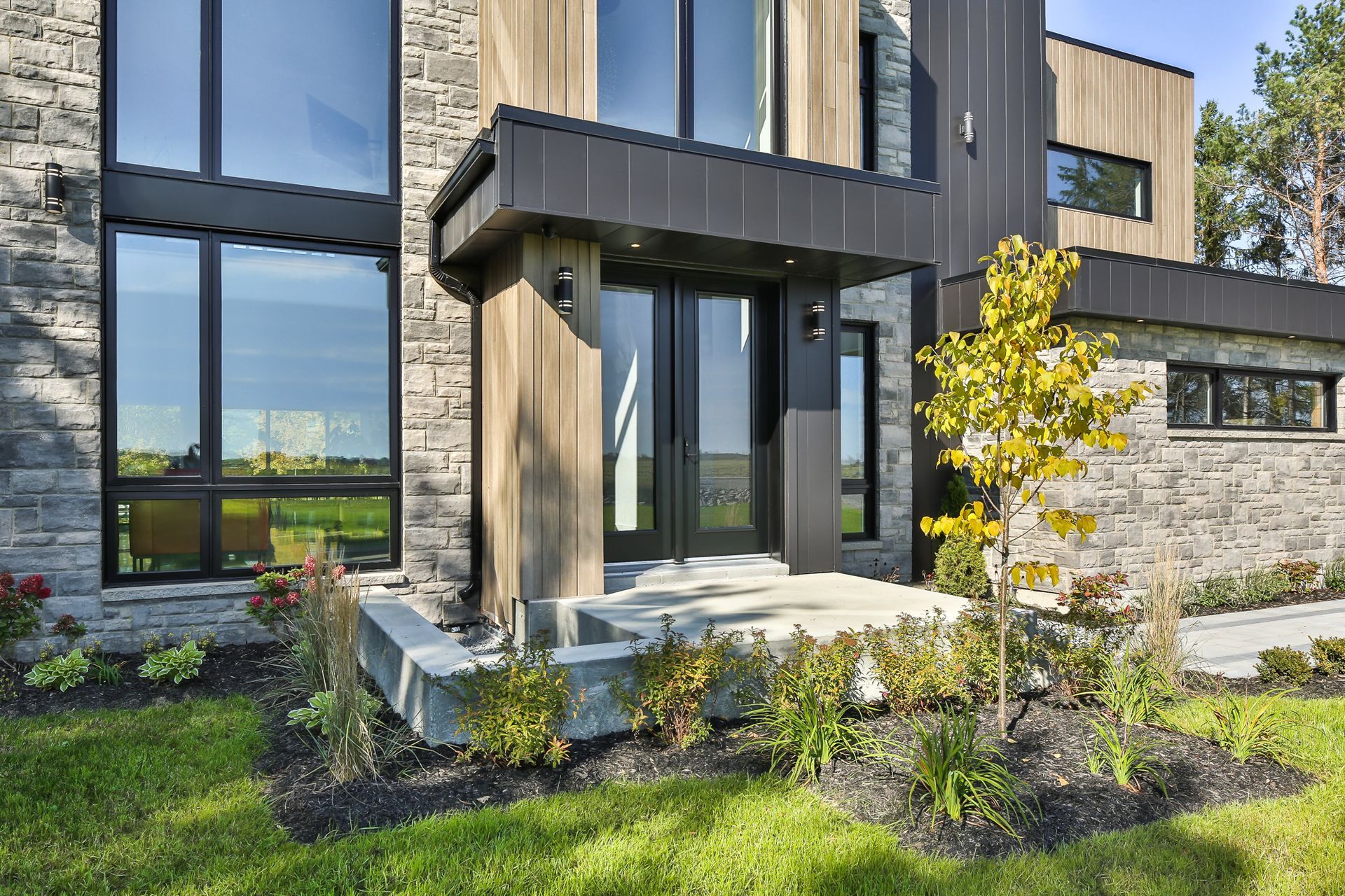Extérieur de maison moderne avec revêtement en pierre et en bois, grandes fenêtres et entrée couverte.