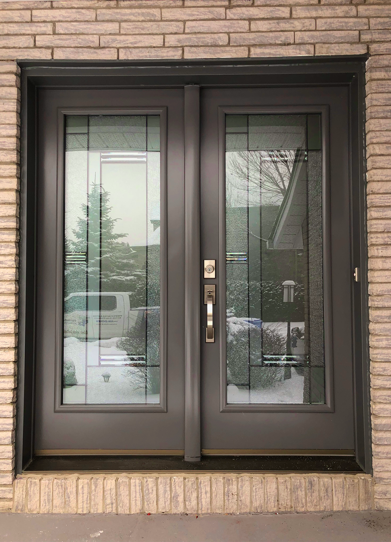 Portes doubles grises avec panneaux de verre et fenêtres latérales dans une façade en brique, recouverte de gouttes d'eau.