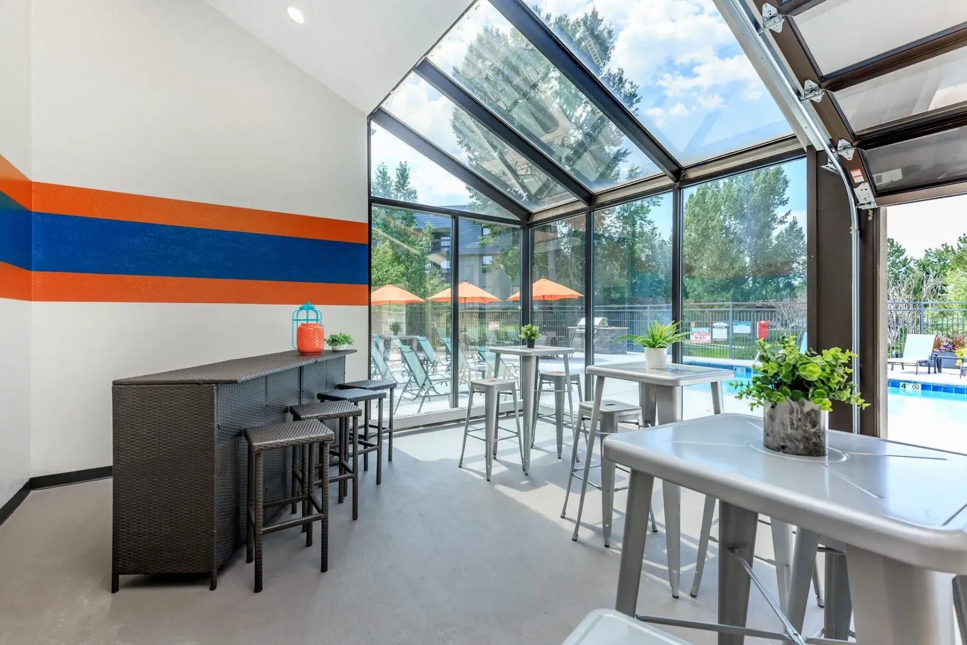 Modern cafe seating area with glass ceiling, black tables, orange-blue wall stripe, and bright outdoor view