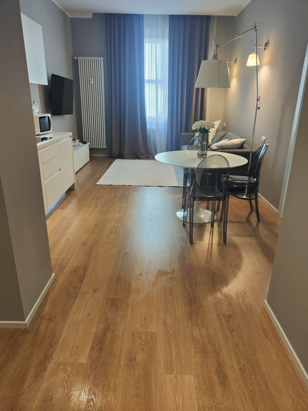 Piccola zona soggiorno con pavimenti in legno chiaro, un tavolo rotondo e un divano vicino a una finestra con tende blu.