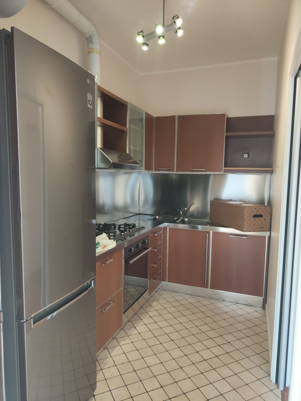 Piccola cucina con mobili marroni, elettrodomestici in acciaio inossidabile e pavimento piastrellato.