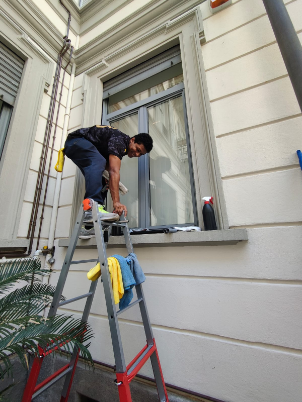 Persona che pulisce una finestra da una scala, accanto a un edificio. Panni gialli e blu sulla scala.