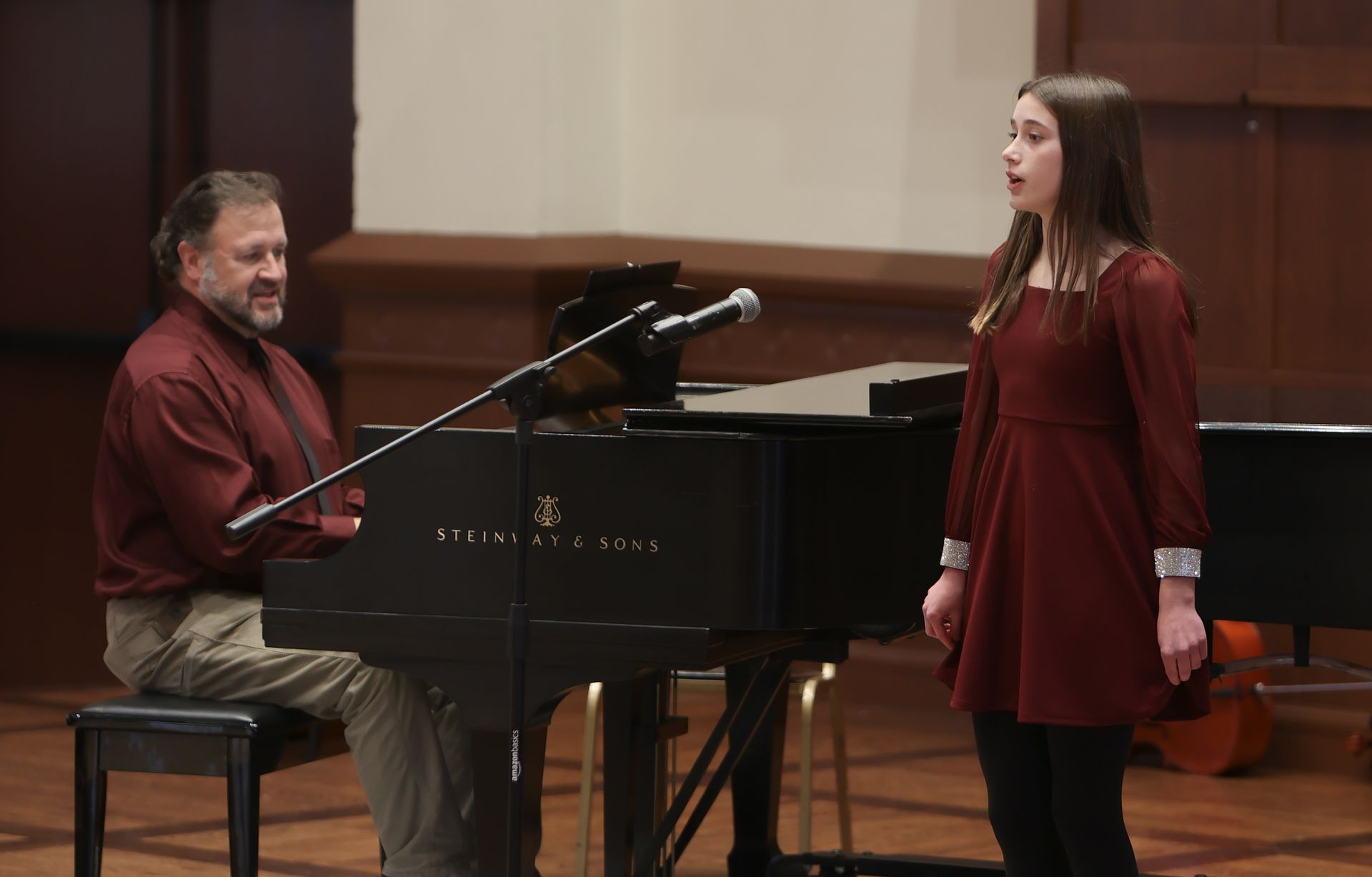 A cello and a smaller viola, both on stands, stand against a wall with a window.