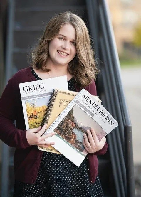Blonde woman in a light pink dress smiles beside a piano in a wood-paneled room.